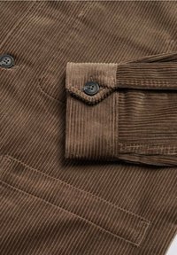 Manica di giacca in corduroy marrone con polsino a bottone nero e bottoni neri abbinati sul tessuto principale, che mostra coste verticali testurizzate.