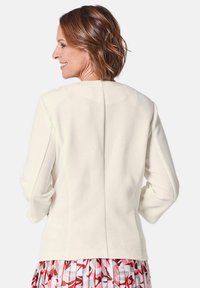 Crème-kleurige textuurjas met lange mouwen, met een centrale naad over de rug. Draag het over een gemusterde rok.