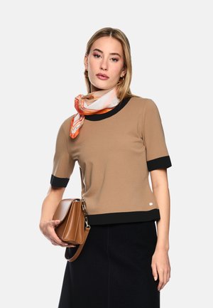 Frau trägt ein hellbraunes Kurzarm-Top mit schwarzem Besatz, ein orange gemustertes Halstuch, einen schwarzen Rock und hält eine braune Lederhandtasche.