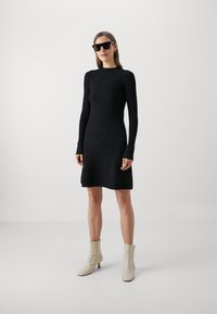 Svart langermet kjole i en tettsittende design, laget av ribbemateriale, kombinert med lyse ankelstøvler og firkantede solbriller.