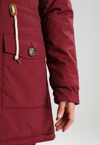 Chaqueta de invierno burdeos con tejido texturizado, bolsillo frontal con botón, cierre con cordón y un ajuste relajado.