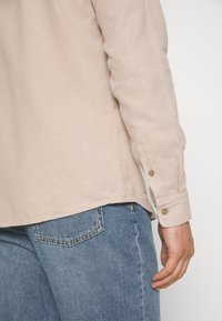 Beżowa koszula z długim rękawem z teksturowanej tkaniny, z zapinanymi mankietami i zaokrąglonym dołem, w zestawieniu z jasnoniebieskimi dżinsami z tylną kieszenią.