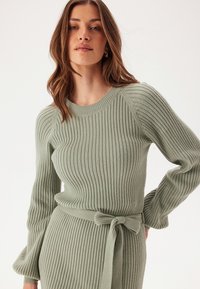 Maglione a coste verde chiaro con scollatura tonda, maniche lunghe e cinturino da annodare in vita. La texture è morbida e leggermente elasticizzata.