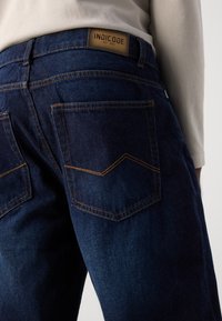 Tumed sinised denim püksid siledat tekstuuri. Omavad kahte tagumist taskut kontrastse õmblusega ning nahast logo sildiga, millel on kirjutatud "INDICODE."