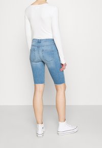 Bermudas de mezclilla azul claro con dobladillo deshilachado, dos bolsillos traseros y un ajuste de tiro medio, combinadas con una camiseta de manga larga blanca y zapatillas blancas.
