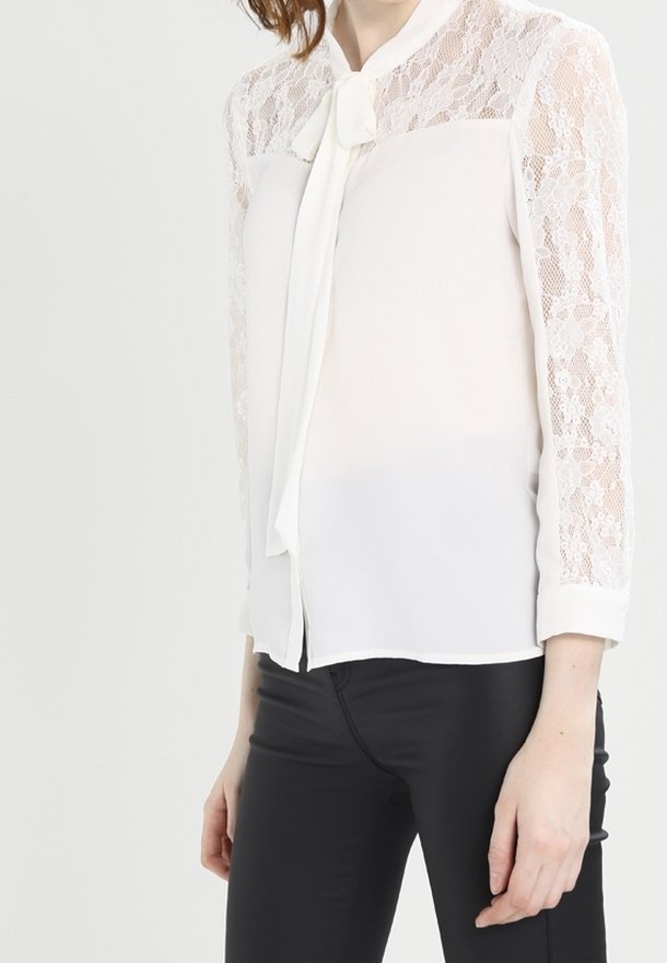 Blusa bianca con yoke in pizzo, maniche lunghe in pizzo e chiusura con fiocco anteriore. Tessuto morbido con una qualità leggera e semi-trasparente.