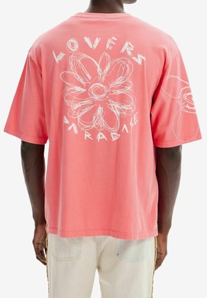 Homme portant un t-shirt surdimensionné corail avec une fleur dessinée en style esquisse blanche et le texte "Lovers Paradise" au dos, associé à un pantalon de couleur claire.