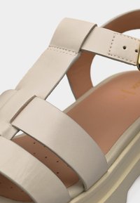 Κοντινό πλάνο μιας μπεζ δερμάτινης σάνδαλας με φαρδιές λωρίδες, χρυσή αγκράφα και επένδυση εσωτερικού πέλματος για άνεση.