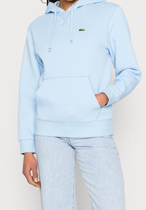 Jasnoniebieska bluza z kapturem z przednią kieszenią i logo Lacoste, noszona z jasnymi dżinsami, ręka w kieszeni, styl casual.
