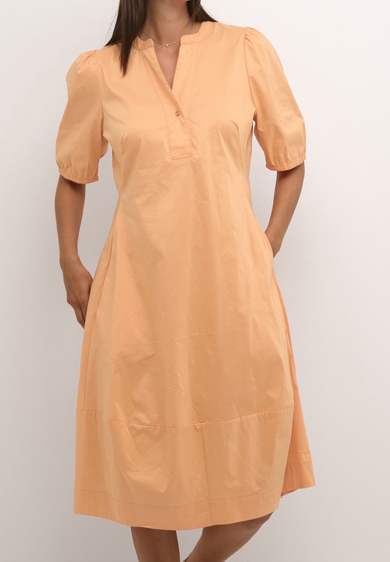 Robe couleur pêche avec manches courtes bouffantes, col à encoche et patte de boutonnage. Elle présente une coupe évasée longueur genoux et des poches latérales.