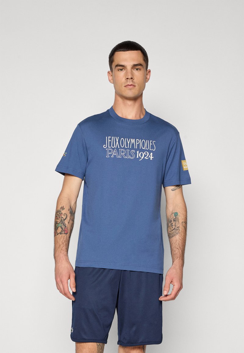 T-shirt en coton bleu avec "JEUX OLYMPIQUES PARIS 1924" imprimé en blanc. Design à manches courtes et col rond, porté avec un short bleu marine.