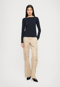 Femeie stând cu mâinile în buzunarele pantalonilor cargo bej, purtând un top cu mânecă lungă bleumarin și tocuri negre cu vârf ascuțit, pe un fundal alb simplu.
