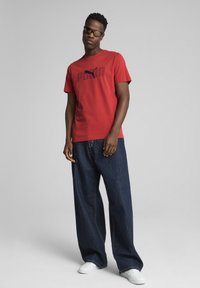 Rød bomulds t-shirt med et sort Puma-logo, parret med løstsiddende mørkeblå denimjeans og hvide sneakers.
