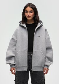 Felpa grigia con zip realizzata in tessuto morbido, dotata di un ampio cappuccio, tasche frontali e un logo ricamato sul petto.