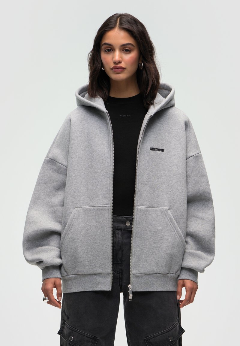 Felpa grigia con zip realizzata in tessuto morbido, dotata di un ampio cappuccio, tasche frontali e un logo ricamato sul petto.