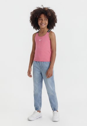 Fille souriante aux cheveux bouclés portant un débardeur Nike rose, un jean jogging bleu clair et des baskets blanches, debout devant un fond blanc uni.