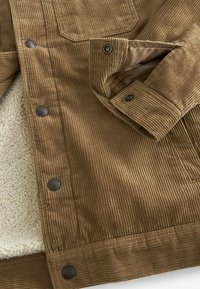 Veste en velours côtelé marron avec boutons-pression métalliques, détail aux poignets et doublure beige en sherpa partiellement visible à l'intérieur.
