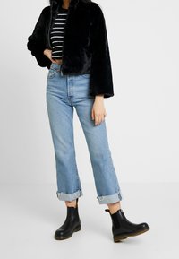 Chaqueta corta de piel sintética negra, camiseta a rayas en blanco y negro, vaqueros de mezclilla azul claro con puños deshilachados y botas Chelsea negras con suelas marrones.