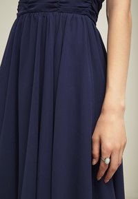 Dłoń kobiety z srebrnym pierścionkiem z białym owalnym kamieniem, leżąca obok granatowej sukienki z marszczonym pasem.