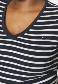 Nærbillede af en person iført en navy- og hvidstribet V-hals t-shirt med et lille rødt, hvidt og blåt logo på brystet.