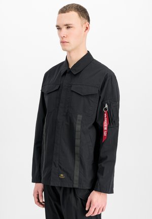 Junger Mann trägt eine schwarze Langarmjacke mit Brusttaschen und einem roten "Remove Before Flight" Anhänger an der Reißverschlusstasche am linken Ärmel.