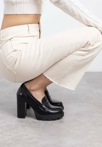 Mocassini neri con plateau dal finish in pelle liscia, tacco largo e suole textured, abbinati a pantaloni cropped di colore chiaro.