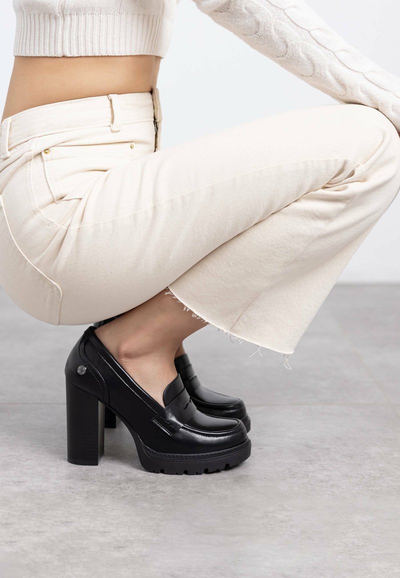 Mocasines de plataforma negros con un acabado de cuero suave, tacones gruesos y suelas texturizadas, combinados con pantalones cortos de color claro.