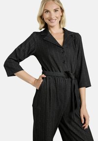 Mustapilkkuinen haalaripuku, jossa on kaulus, napitettu etuosa ja kolme neljännestä hihoista. Varustettu vyötärön vyöllä ja tekstuurilla.