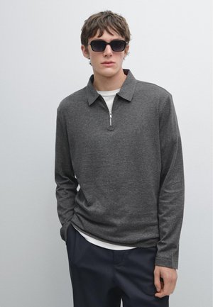 Donkergrijze longsleeve shirt met een ritskraag, gelaagd over een wit shirt. Model draagt zwarte zonnebril en marineblauwe broek. Eenvoudige textuur, casual ontwerp.