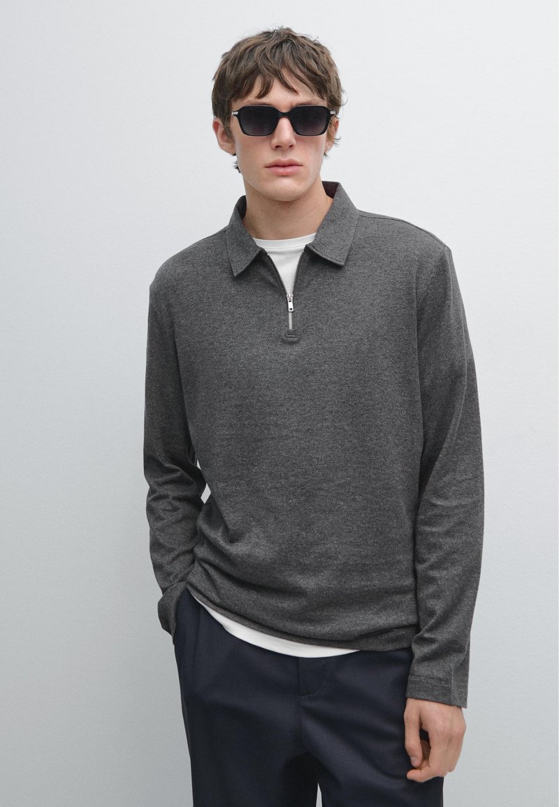 Dunkelgraues Langarmshirt mit einem Reißverschlusskragen, über einem weißen Shirt getragen. Das Modell trägt schwarze Sonnenbrillen und marineblaue Hosen. Einfache Textur, lässiges Design.