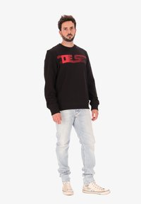 Uomo in piedi che indossa una felpa nera con scritte rosse, jeans chiari e scarpe alte color avorio su uno sfondo bianco.