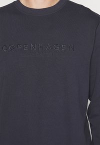 Camiseta de manga larga azul marino con el texto bordado "COPENHAGEN LINDBERGH" en el pecho. Tela suave, cuello redondo y ajuste clásico.