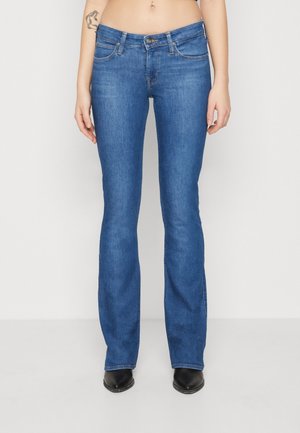 Femme portant un jean bootcut bleu et des bottes pointues noires, debout devant un fond blanc uni avec un tatouage visible sur son bras gauche.