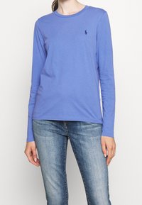 T-shirt à manches longues en coton bleu clair avec un col rond et un petit logo bleu marine sur la poitrine gauche, associé à un jean en denim bleu.