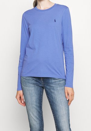 T-shirt à manches longues en coton bleu clair avec un col rond et un petit logo bleu marine sur la poitrine gauche, associé à un jean en denim bleu.