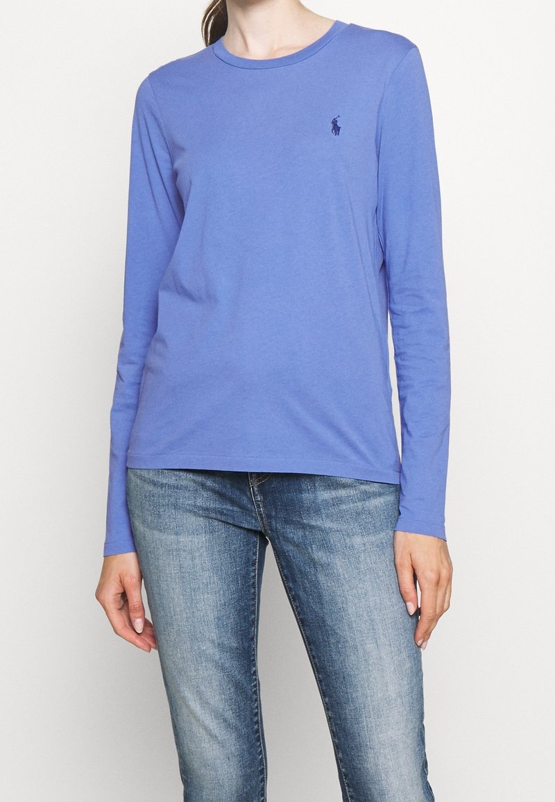 T-shirt à manches longues en coton bleu clair avec un col rond et un petit logo bleu marine sur la poitrine gauche, associé à un jean en denim bleu.