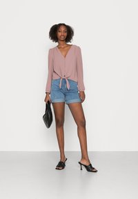 Light pink long-sleeve blouse with a tie at the waist, paired with denim shorts. Accessories include a black woven handbag and heeled sandals.