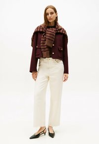 Bordeaux cropped jasje met gouden knopen, beige wijde broek met franjes aan de onderkant, en een gestreepte bruine sjaal om de hals. Zwarte slingback schoenen.