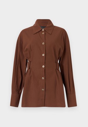 Chemise marron à manches longues avec boutons sur le devant, col large et détails de boucles latérales réglables, présentée sur un fond uni.