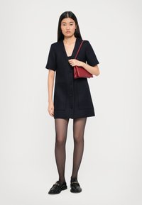 Fiatal nő fekete gombos ruhát, áttetsző fekete harisnyát, fekete lakkcipőt visel, és egy kis piros válltáskát tart, amelynek vállpántja van.