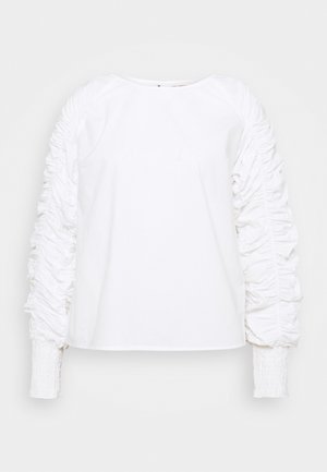 Blouse blanche à manches longues avec manches froncées et resserrées aux poignets par un effet smocké, dotée d'un col rond et d'une coupe ample.