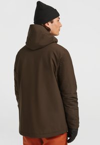 Bruine geïsoleerde jas met een capuchon, met een gladde textuur. Gedragen over oranje broeken, aangevuld met een zwarte gebreide muts en handschoenen.
