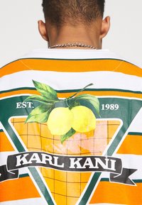 Pruhovaná krátká košile s krátkým rukávem v bílé, oranžové a zelené barvě s trojúhelníkovou grafikou, která zobrazuje citrony a mřížkovaný vzor, logo "KARL KANI".