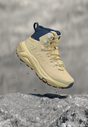 Botte de randonnée beige et marine avec semelle Vibram en l'air au-dessus d'un sol rocheux, montrant un mouvement avec de la terre qui tombe de la semelle.