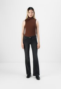 Bruine mouwloze coltrui gecombineerd met zwarte flare jeans. Glad materiaal, slim fit ontwerp en zilveren knoopdetails op de jeans.