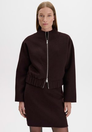 Donna con capelli lisci indossa giacca marrone scuro con zip e gonna abbinata, in piedi davanti a uno sfondo chiaro e uniforme.
