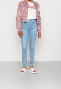 Ljusrosa bomberjacka, vit t-shirt med grafik, ljusblå tajta jeans och vita sandaler med korsremmar. Släta texturer.