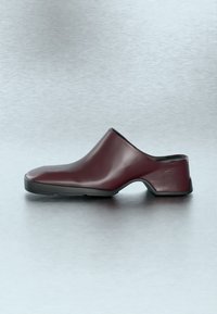 Μπορντό δερμάτινο παπούτσι χωρίς κορδόνια με χαμηλό τακούνι και μαύρη σόλα, σε φόντο με λείο, ανοιχτό γκρι χρώμα.