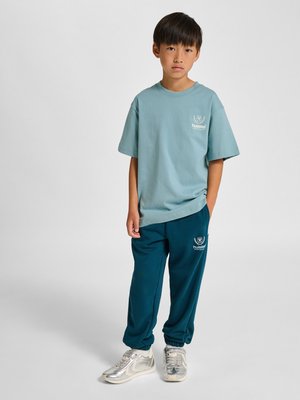 Junger Junge steht, trägt hellblaues T-Shirt und dunkelblaue Jogginghose mit weißen Turnschuhen, neutraler Hintergrund.