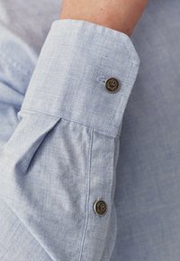 Manșeta cămășii de culoare albastră deschisă, prezentând un material texturat, un design pliat și un buton decorativ închis la culoare cu un logo.